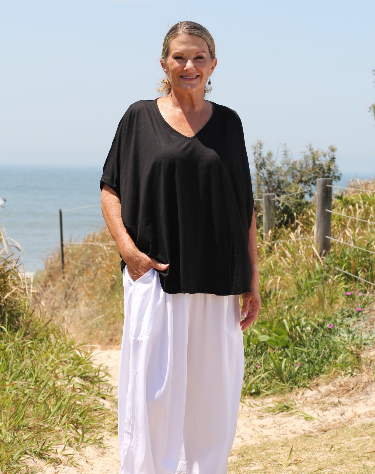 Woman wearing a black comfort tee and white pants standing on a path near the ocean.