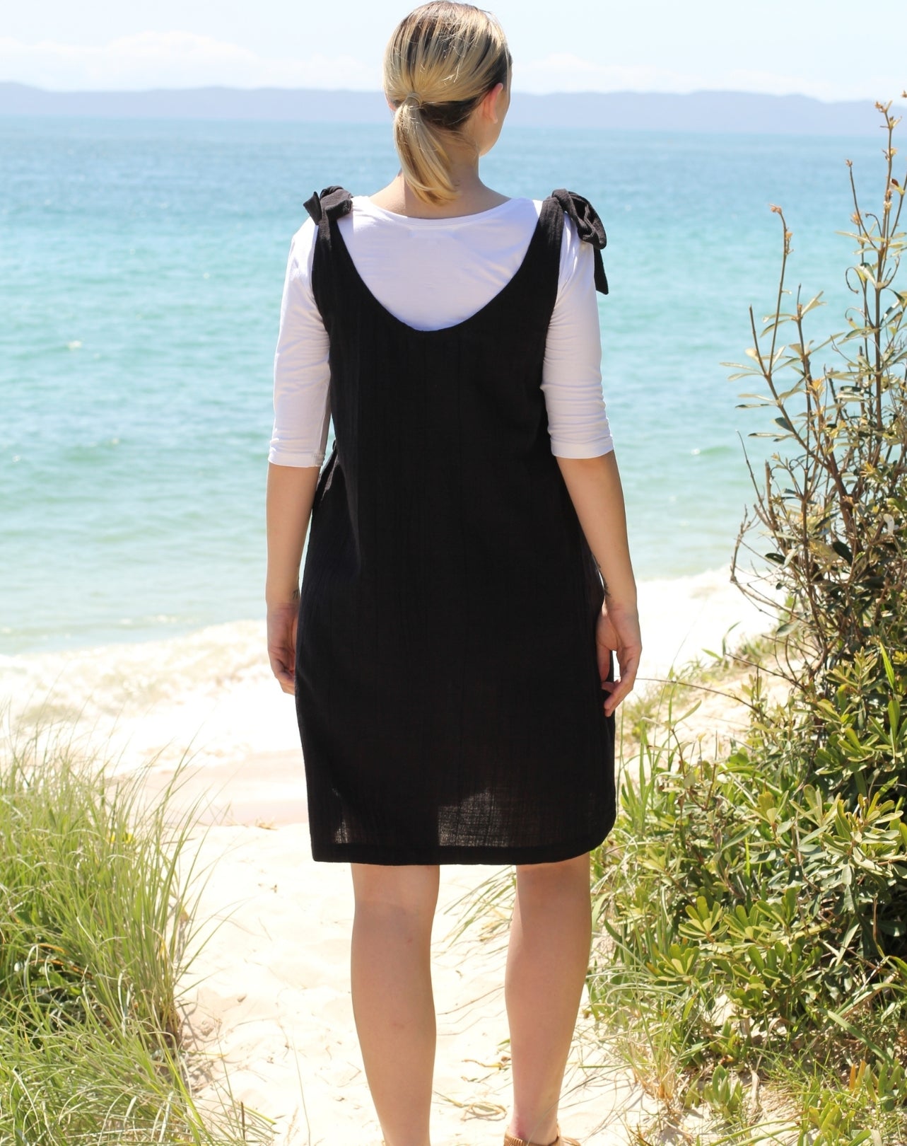 A woman stands on a sandy path by the ocean, viewed from behind, wearing a black sleeveless pinafore-style dress over a white three-quarter-sleeve top.
