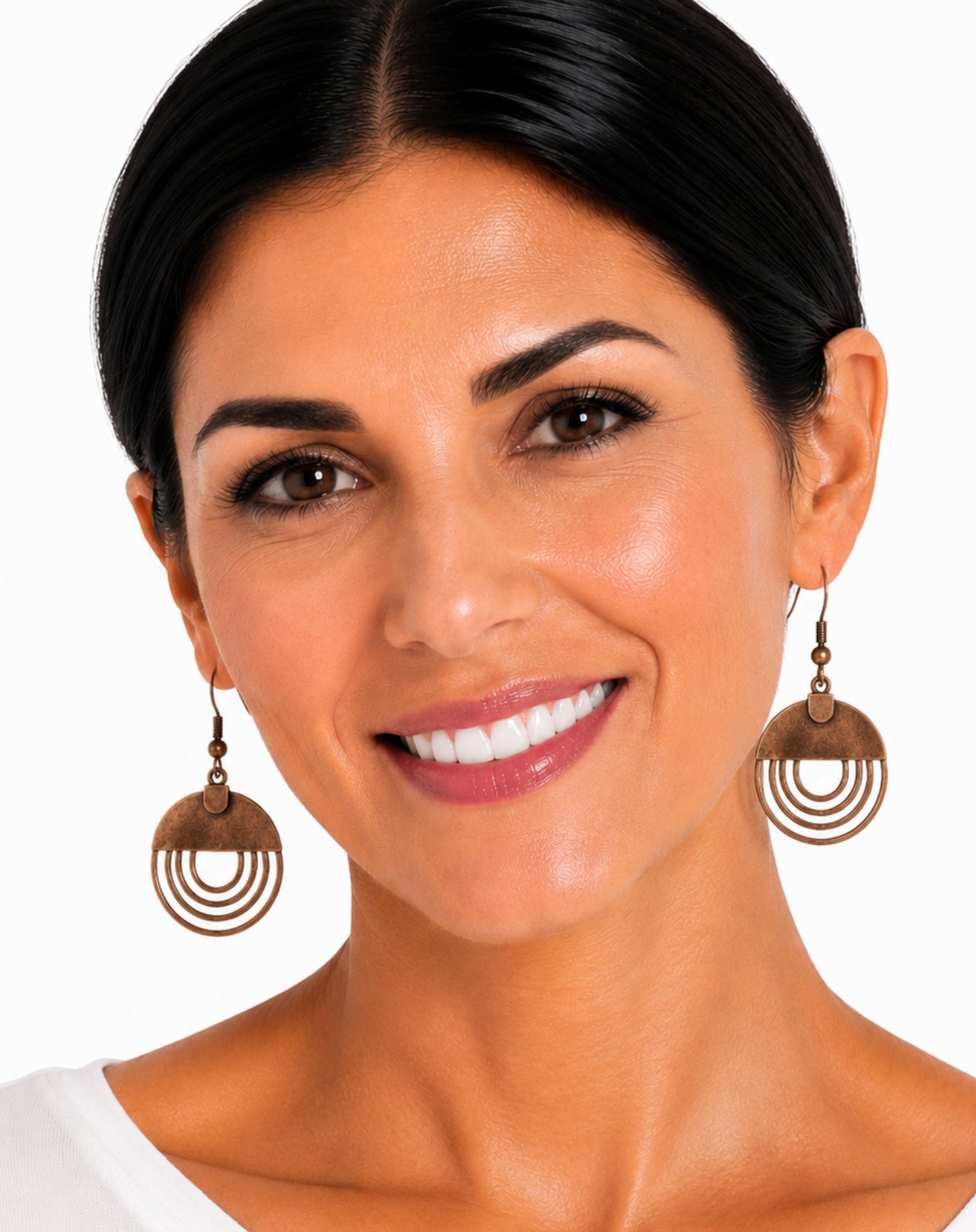 A close-up portrait of a smiling woman with dark hair pulled back, wearing large bronze-toned circular drop earrings against a plain white background.