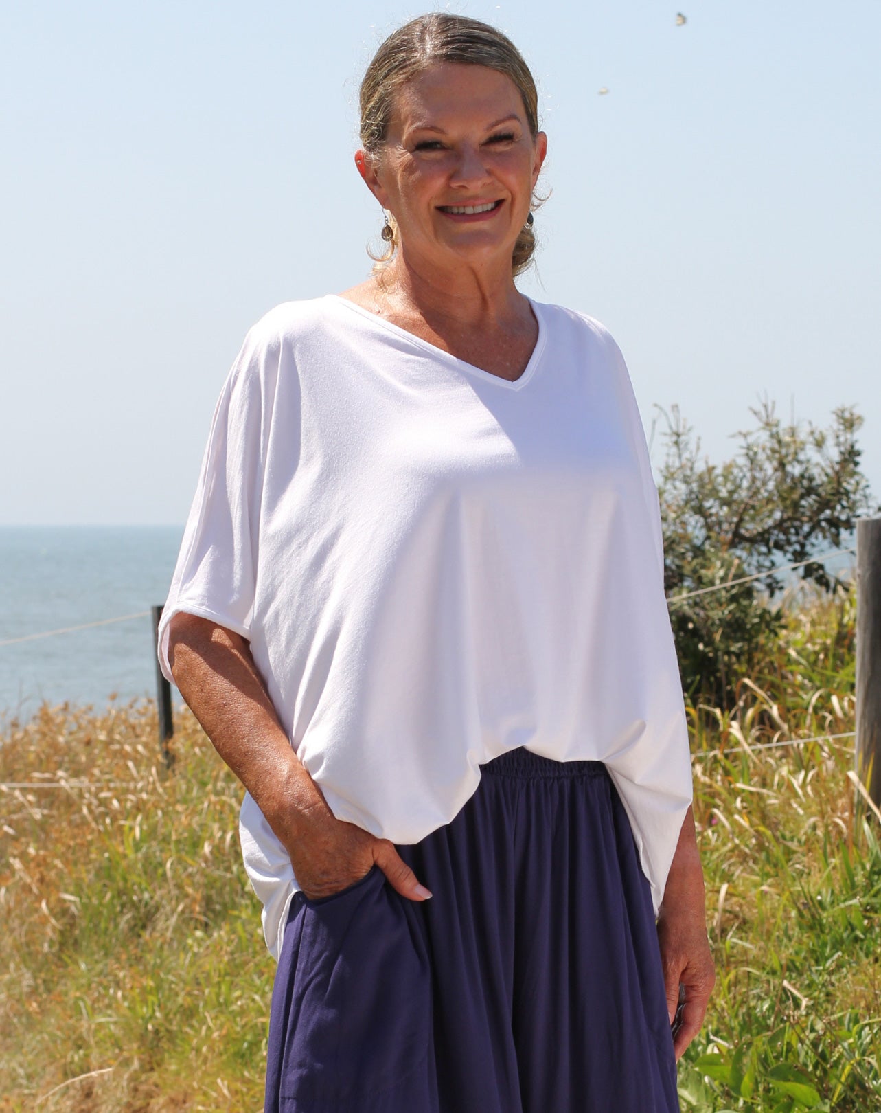 Woman wearing a white tee and dark blue pants standing in a field with a clear sky.