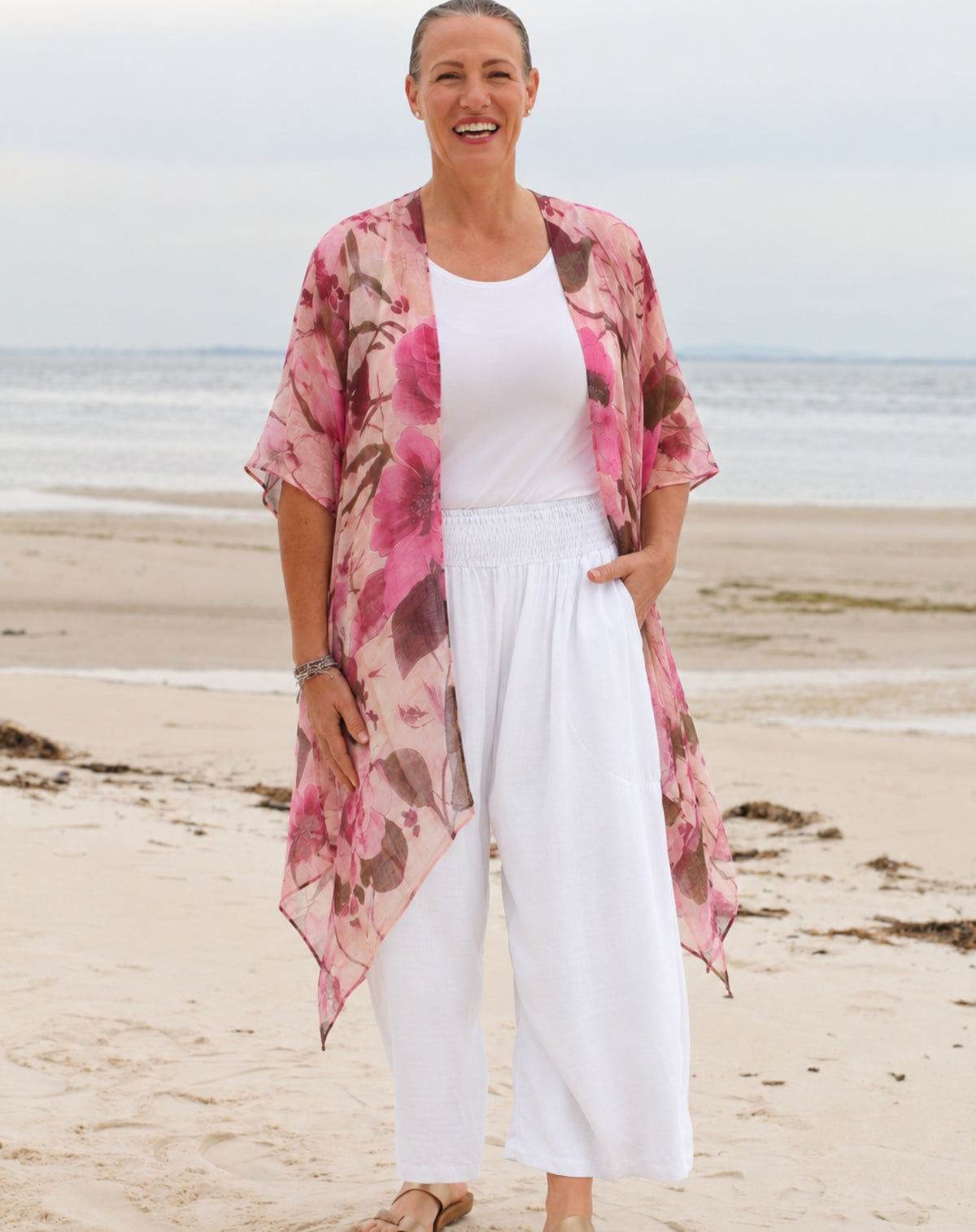 A woman standing on a sandy beach wearing a pink floral kimono cardigan over a white tank top and white wide-leg pants.