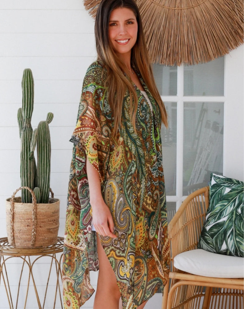 Woman wearing a paisley print in green kimono cover up jacket standing in a room with a boho wicker chair.