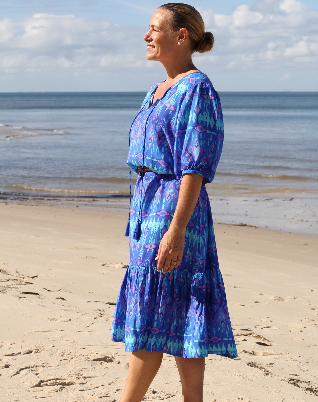 A woman wearing a blue patterned midi dress stands on a sandy beach with the ocean and cloudy sky in the background.