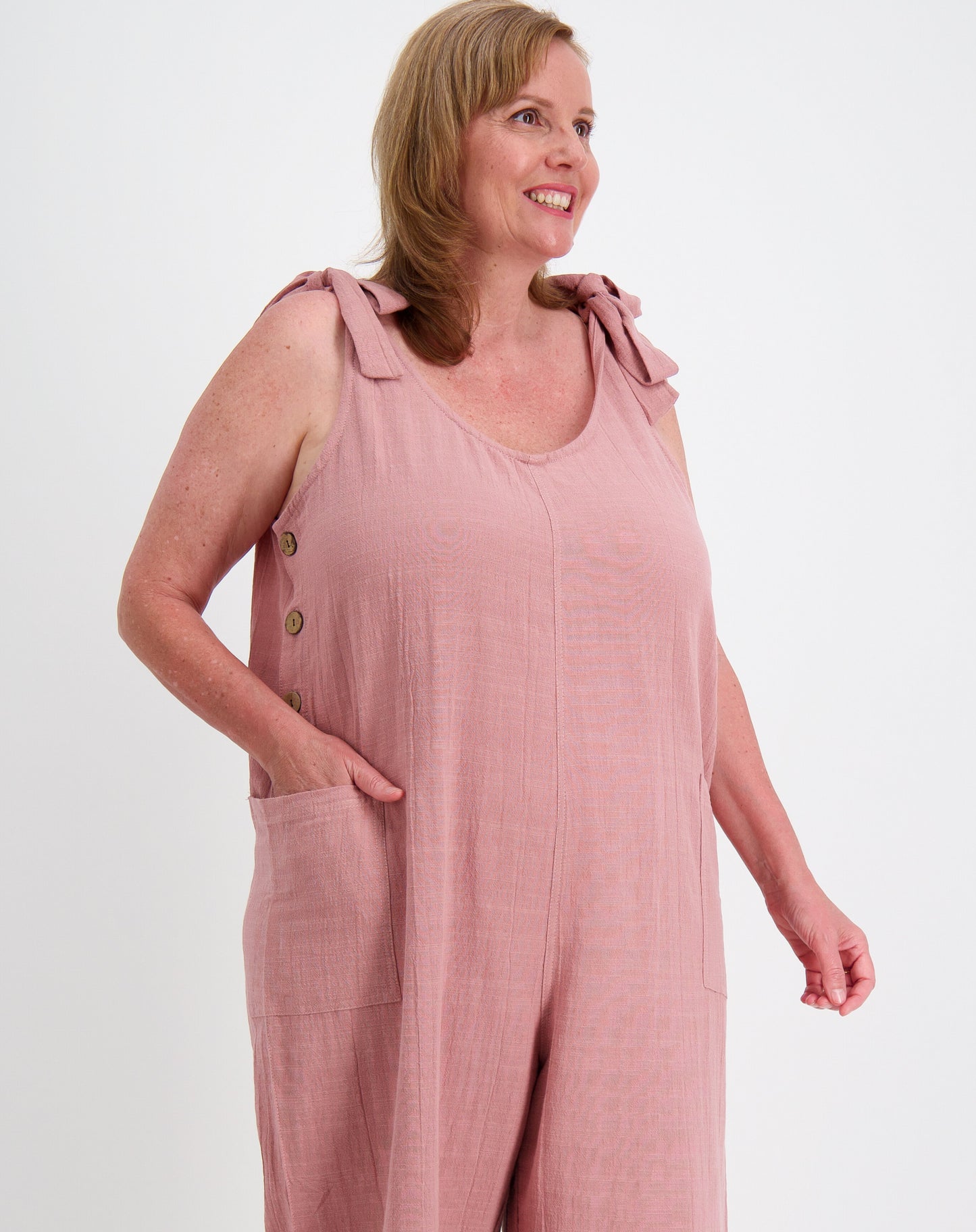 mature woman wearing tiestrap jumpsuit in blush pink with button details on a white background
