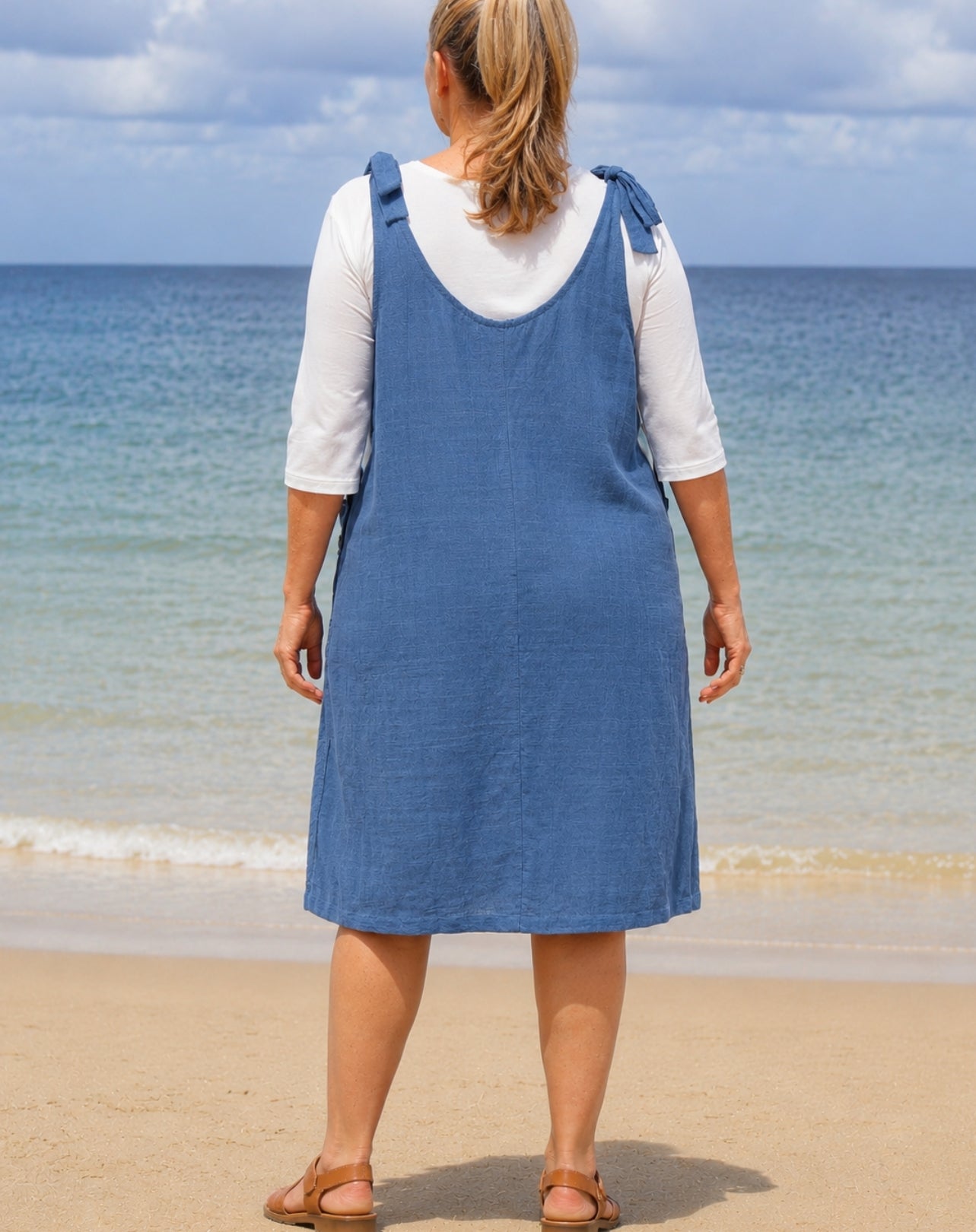 A woman stands on a sandy beach facing the ocean, wearing a blue denim pinafore dress over a white short-sleeve t-shirt and brown sandals.