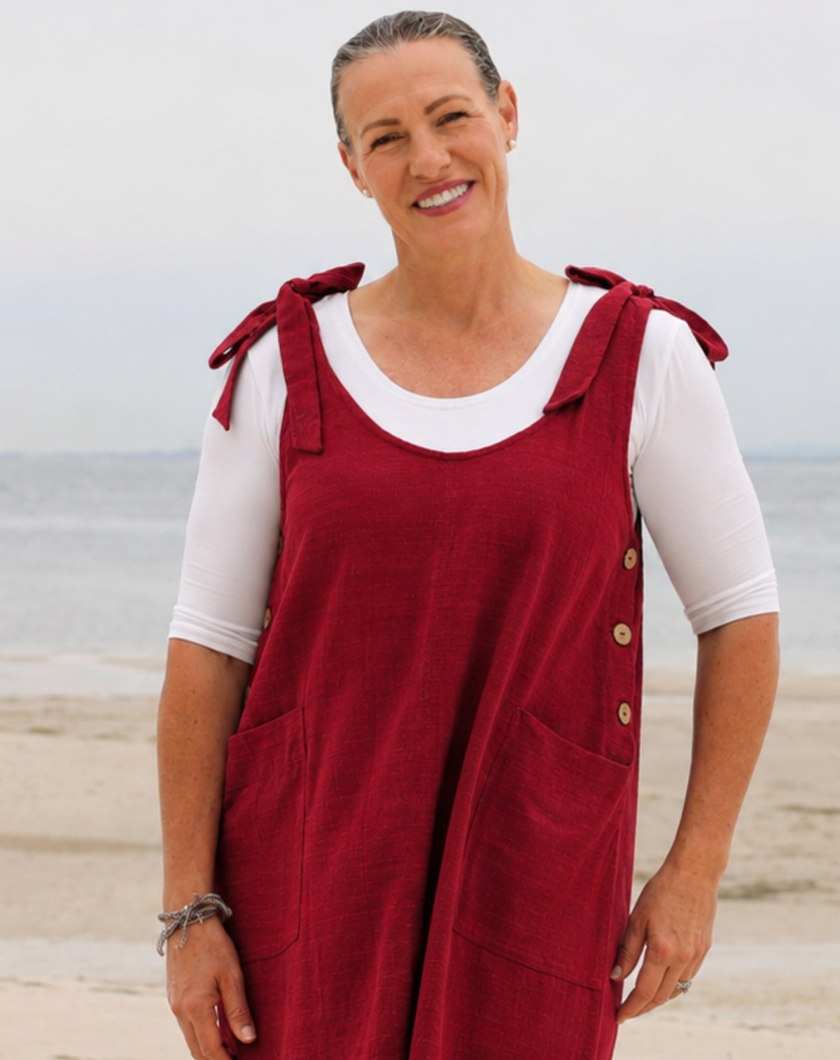 A smiling woman standing on a beach wearing a red linen overall dress over a white short-sleeve t-shirt. The dress features tie straps on the shoulders, button details on the side, and large front patch pockets.