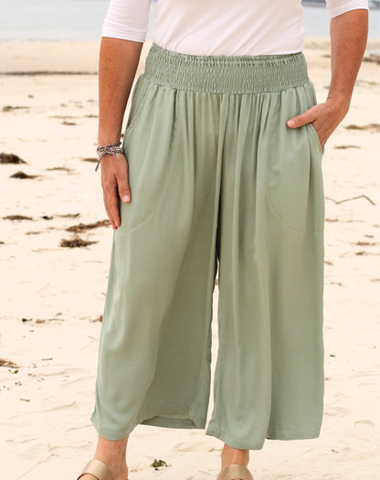 A person standing on a sandy beach wearing light mint green wide-leg cropped pants with an elastic waistband, side pockets, and a white short-sleeve top.