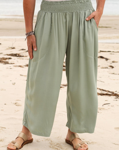 A person standing on a sandy beach wearing loose, wide-leg mint green pants with an elastic waistband and side pockets, paired with tan slide sandals.