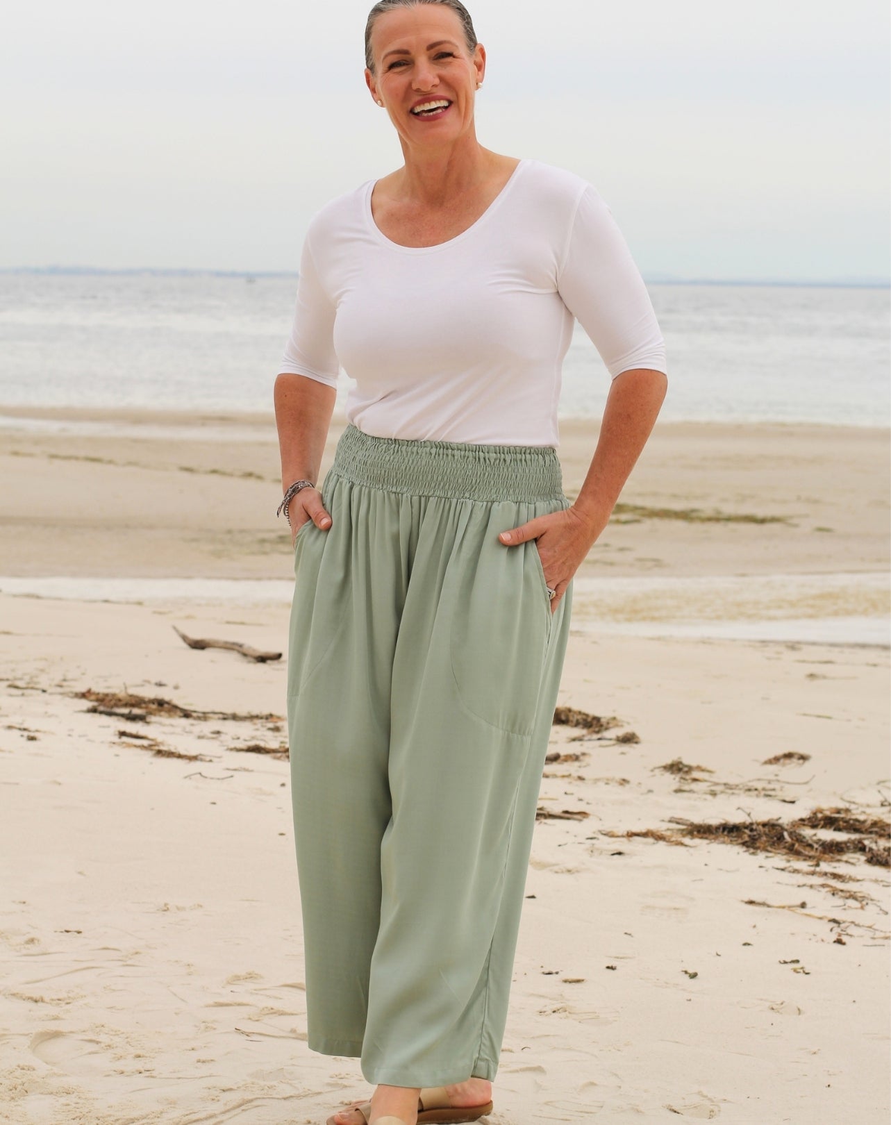 A woman standing on a sandy beach wearing a white short-sleeve scoop-neck top and mint green wide-leg pants with an elastic waistband.