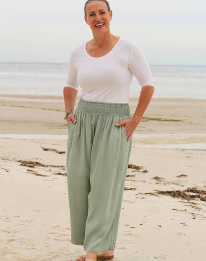 A woman standing on a sandy beach wearing a white short-sleeve scoop-neck top and mint green wide-leg pants with an elastic waistband.