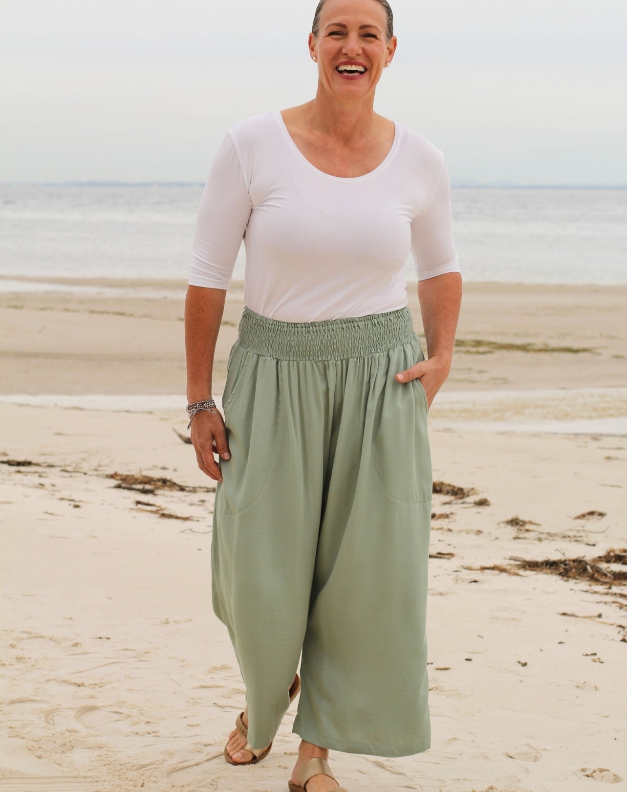 A woman standing on a sandy beach wearing a white short-sleeve scoop-neck top and mint green 7/8 wide-leg pants with an elastic waistband.