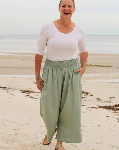 A woman standing on a sandy beach wearing a white short-sleeve scoop-neck top and mint green 7/8 wide-leg pants with an elastic waistband.