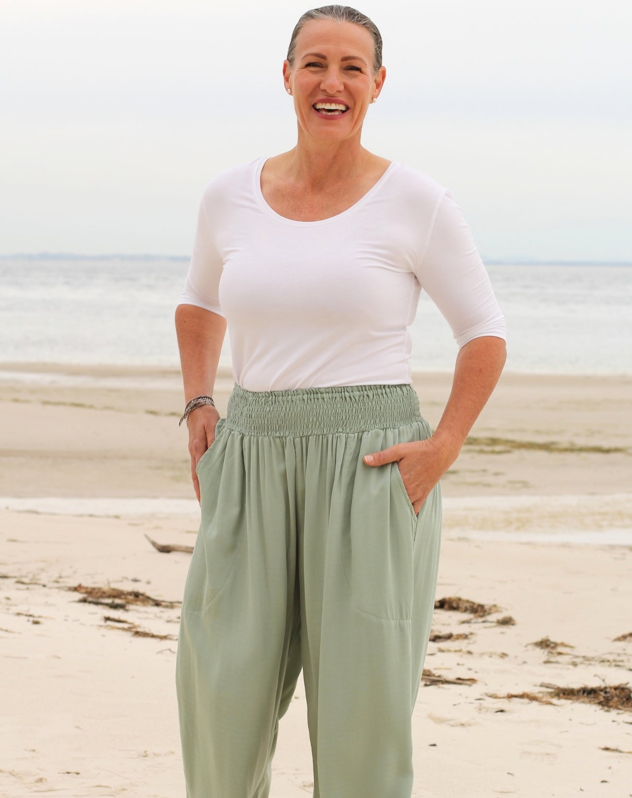 A woman standing on a sandy beach wearing a white short-sleeve scoop-neck top and mint green high-waisted, loose-fit pants with deep side pockets.