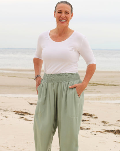 A woman standing on a sandy beach wearing a white short-sleeve scoop-neck top and mint green high-waisted, loose-fit pants with deep side pockets.
