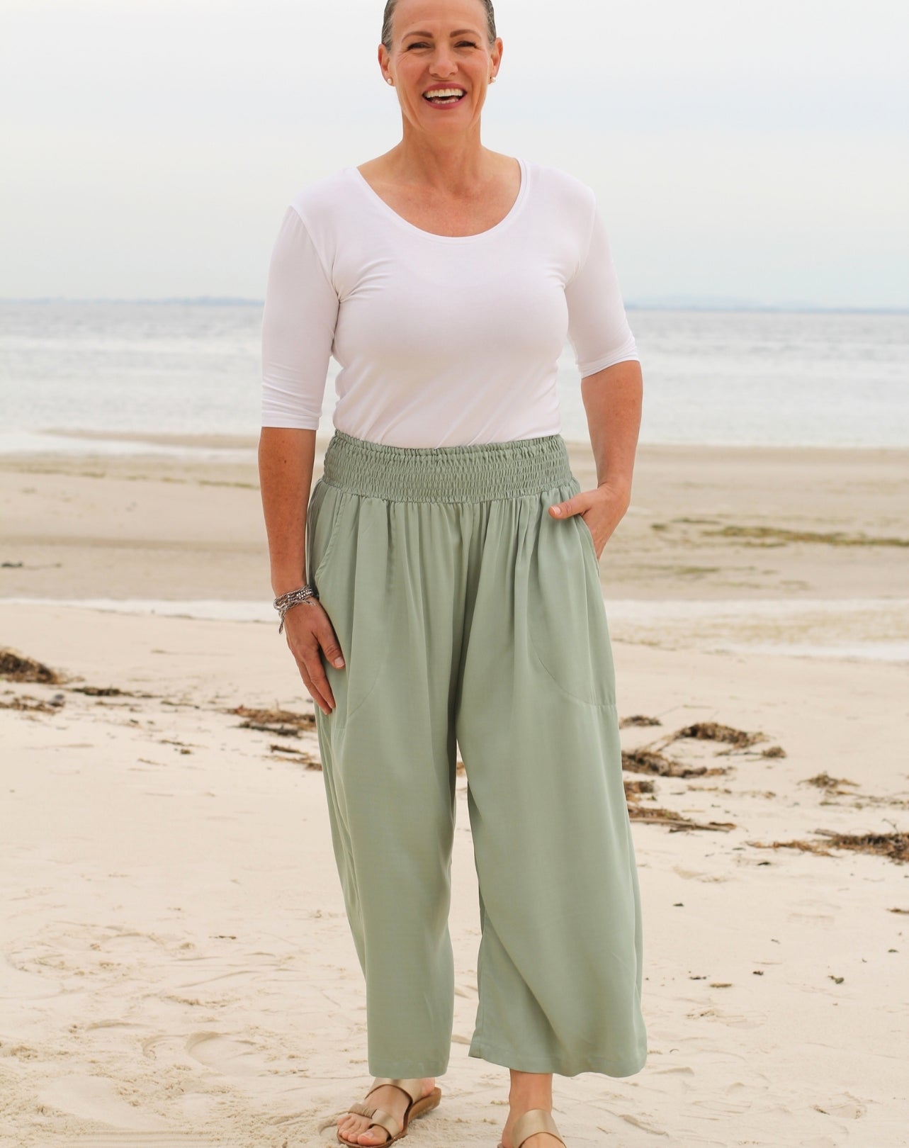 A woman standing on a sandy beach wearing light mint green wide-leg pants 7/8 length with a smocked elastic waistband, paired with a white short-sleeve top.