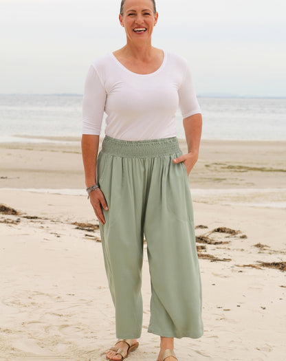 A woman standing on a sandy beach wearing light mint green wide-leg pants 7/8 length with a smocked elastic waistband, paired with a white short-sleeve top.