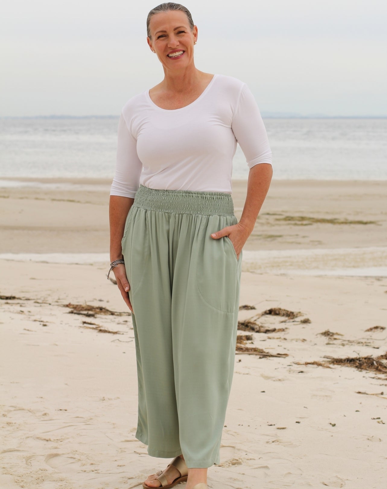 A woman standing on a sandy beach wearing a white short-sleeve top and sage green wide-leg pants.