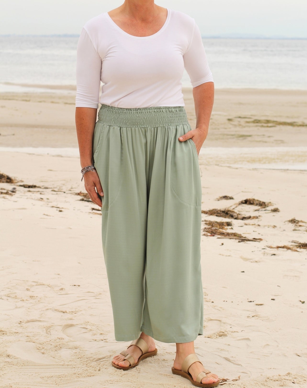 A woman standing on a sandy beach wearing light mint green wide-leg cropped pants with a smocked waistband, a white short-sleeve top, and brown sandals.