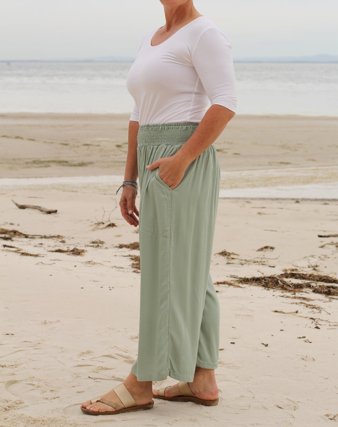 A woman standing on a sandy beach wearing a white short-sleeve top and sage green high-waisted, wide-leg pants with side pockets, paired with beige sandals.