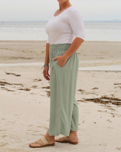 A woman standing on a sandy beach wearing a white short-sleeve top and sage green high-waisted, wide-leg pants with side pockets, paired with beige sandals.