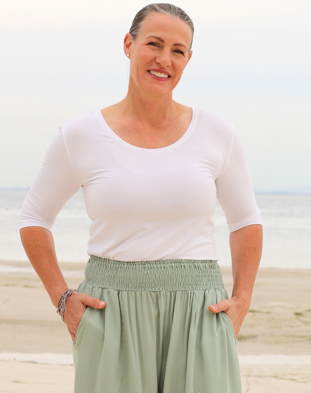 A woman standing on a beach wearing a fitted white scoop-neck short-sleeve top and mint green wide-leg pants with a smocked waistband.