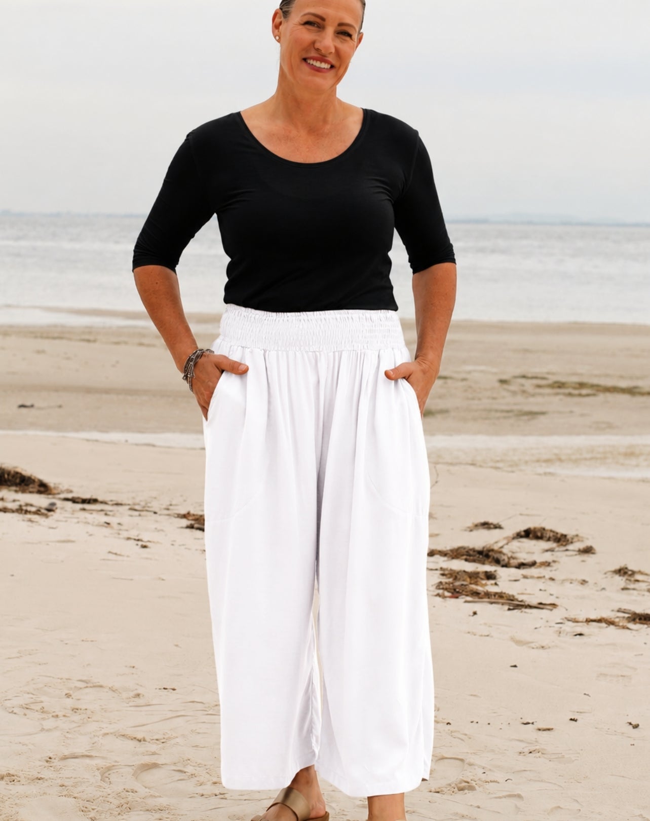 A woman standing on a sandy beach wearing a black short-sleeve scoop-neck top and high-waisted white wide-leg pants with side pockets.