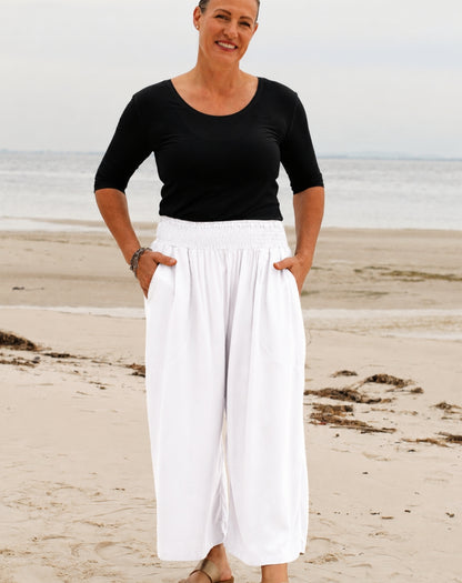 A woman standing on a sandy beach wearing a black short-sleeve scoop-neck top and high-waisted white wide-leg pants with side pockets.