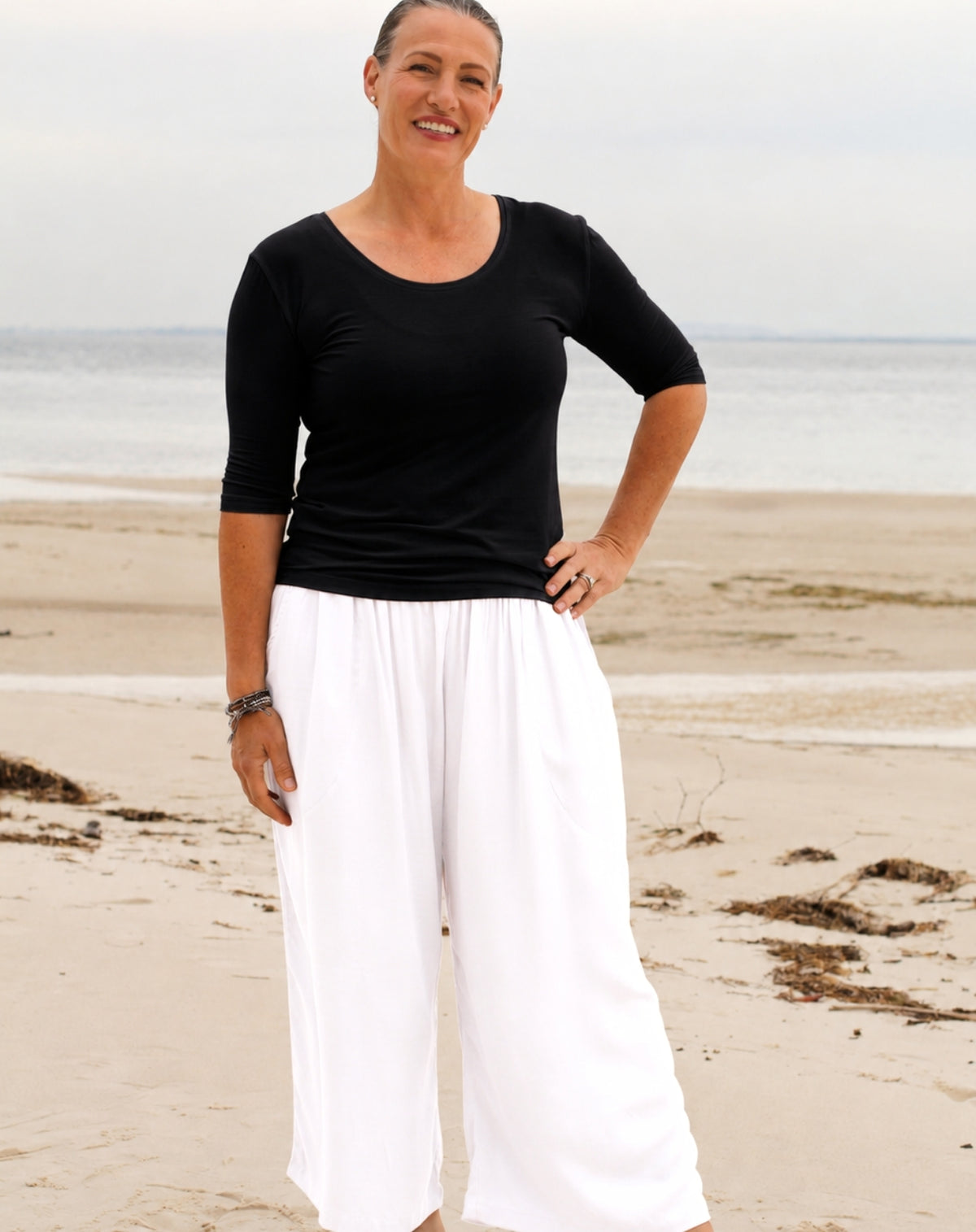 A woman standing on a sandy beach wearing a black three-quarter-sleeve scoop-neck top and white wide-leg pants.