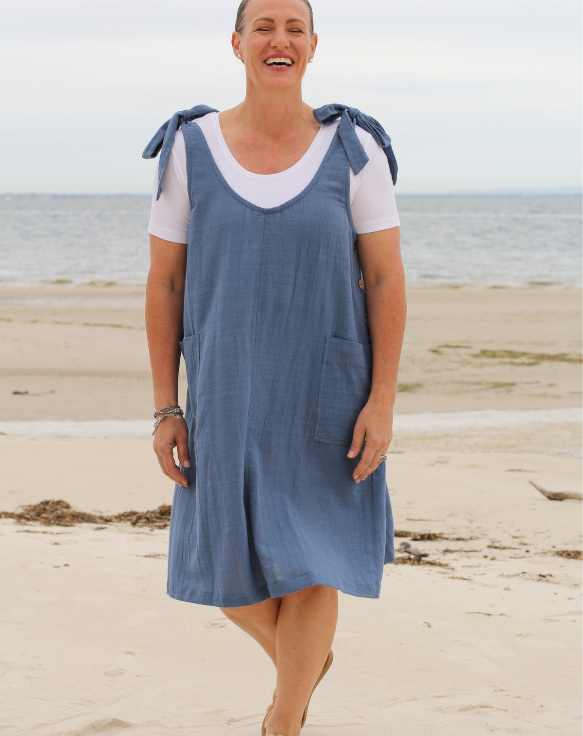 A woman smiling while walking on a sandy beach, wearing a blue linen look pinafore dress over a white t-shirt.