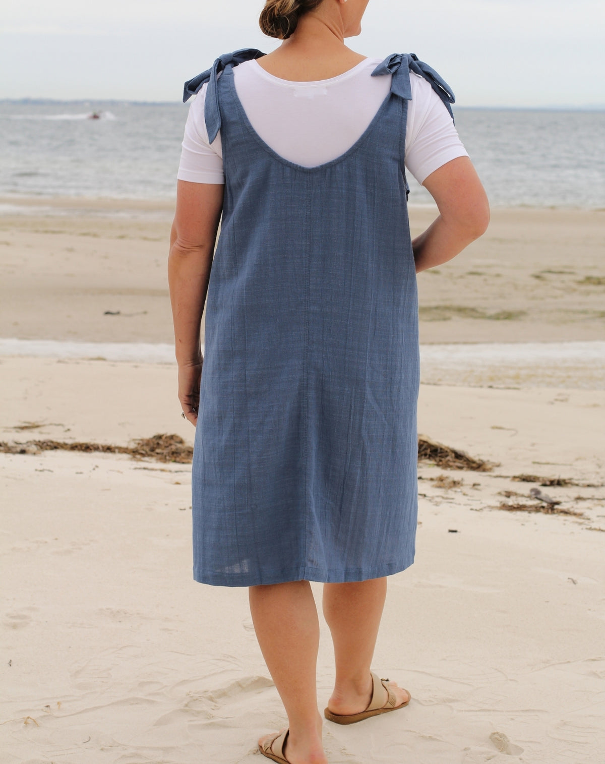 A woman standing on a sandy beach viewed from behind, wearing a blue linen-look pinafore dress over a white t-shirt.