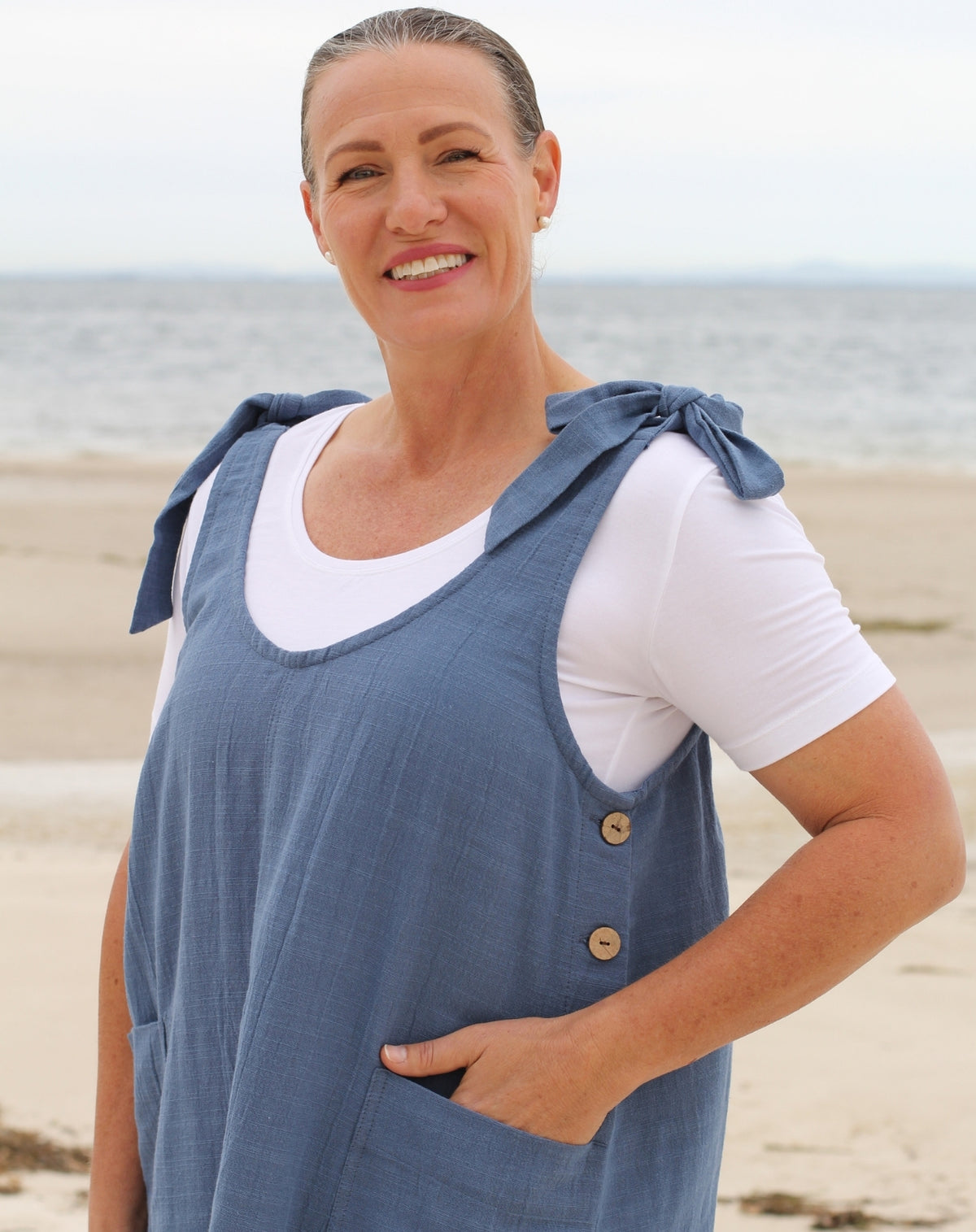 A smiling woman standing on a sandy beach wearing a slate blue cotton linen-look smock dress over a white t-shirt. The dress features tie details on the shoulders and two wooden buttons on the side.