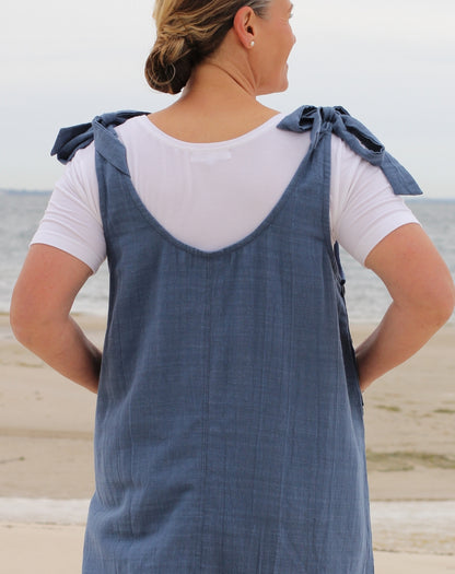 A woman on a beach wearing denim tie strap smock dress over a white t-shirt, viewed from behind to show the back of the dress with its tie shoulder straps.