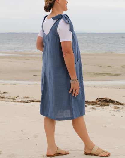 A woman standing on a sandy beach wearing a blue cotton pinafore smock dress over a white t-shirt and tan sandals.