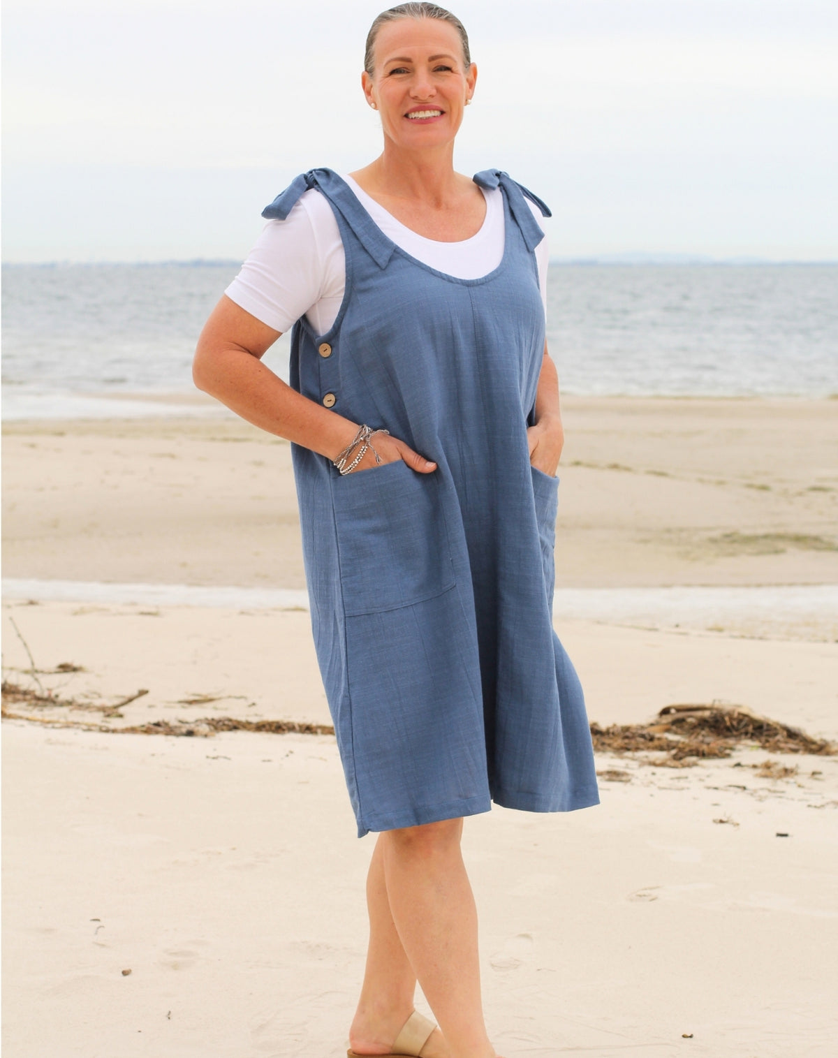 A woman standing on a sandy beach wearing a blue denim tie strap smock dress over a white t-shirt.