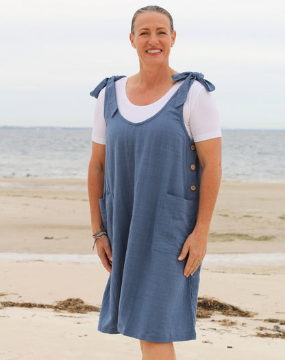 A woman standing on a sandy beach wearing a blue linen-look smock dress over a white t-shirt.