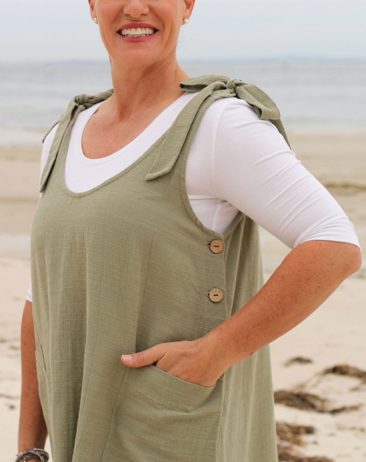 A woman wearing a sage green sleeveless smock-style dress over a white long-sleeve shirt, standing on a sandy beach with the ocean in the background.