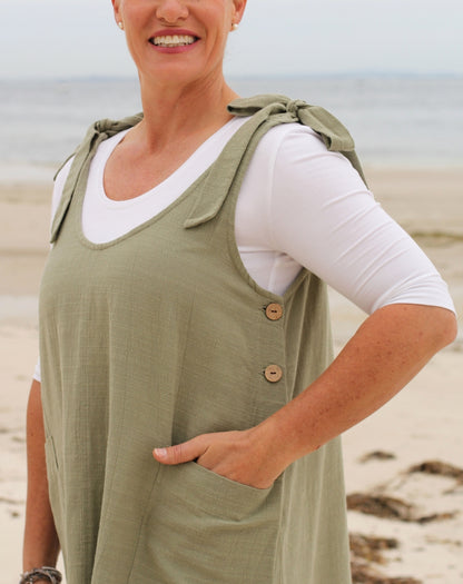 A woman wearing a sage green sleeveless smock-style dress over a white long-sleeve shirt, standing on a sandy beach with the ocean in the background.
