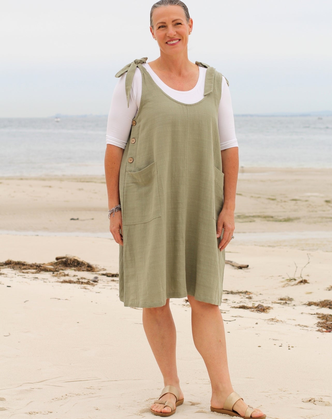 A woman standing on a sandy beach wearing a sage green linen tie strap smock dress over a white t-shirt and tan sandals.