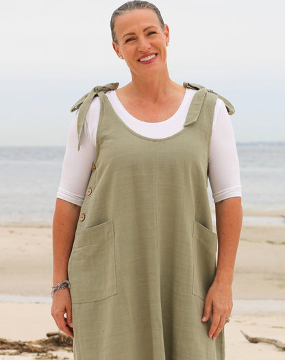 A smiling woman standing on a sandy beach wearing a sage green sleeveless pinafore-style smock dress over a white short-sleeve top. The dress has tie details at the shoulders, decorative buttons along the neckline, and large front patch pockets.