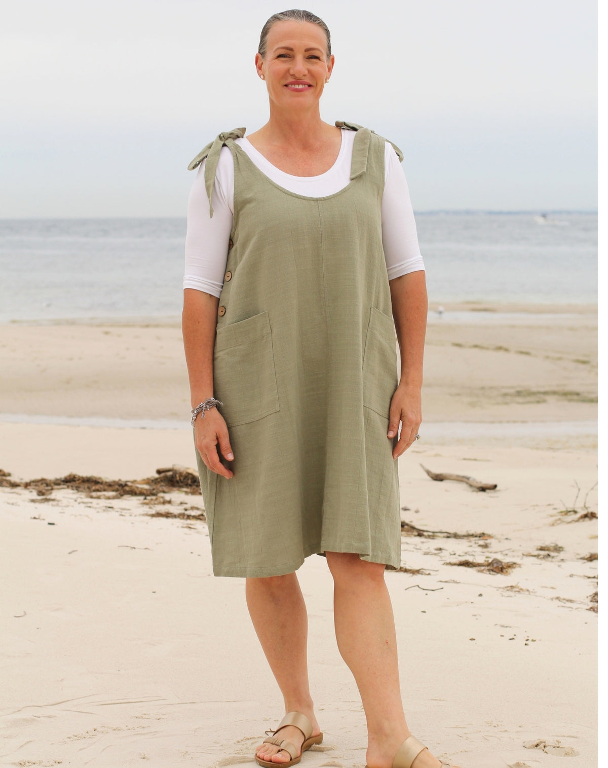A woman standing on a sandy beach wearing a sage green linen pinafore dress over a white short-sleeve t-shirt.