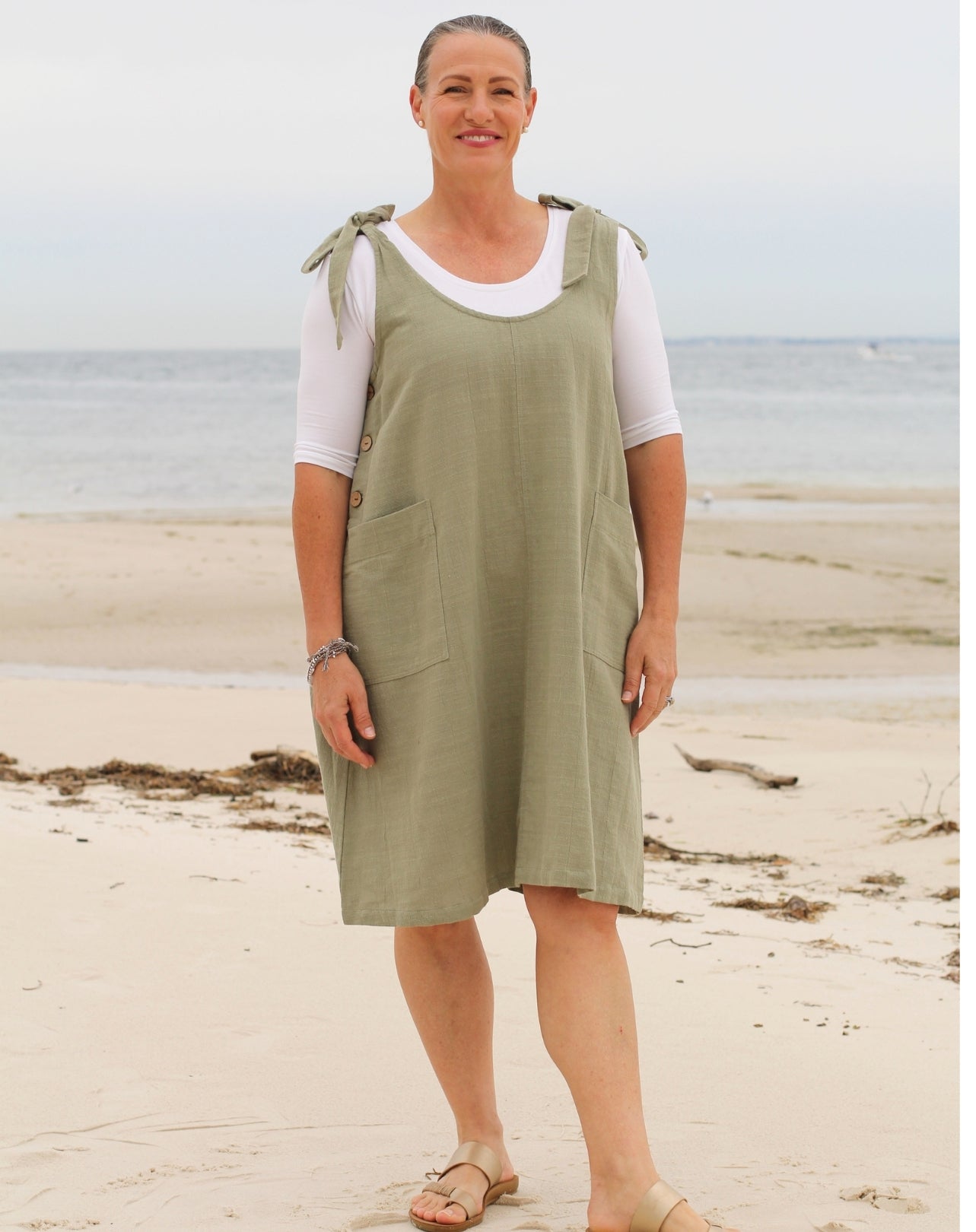 A woman standing on a sandy beach wearing a sage green linen pinafore dress over a white short-sleeve t-shirt.
