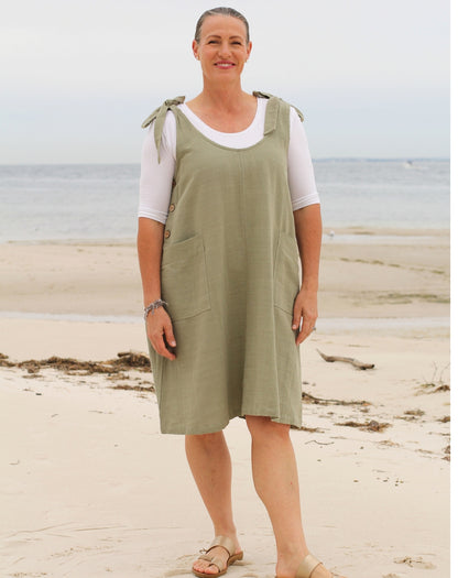 A woman standing on a sandy beach wearing a sage green linen pinafore dress over a white short-sleeve t-shirt.