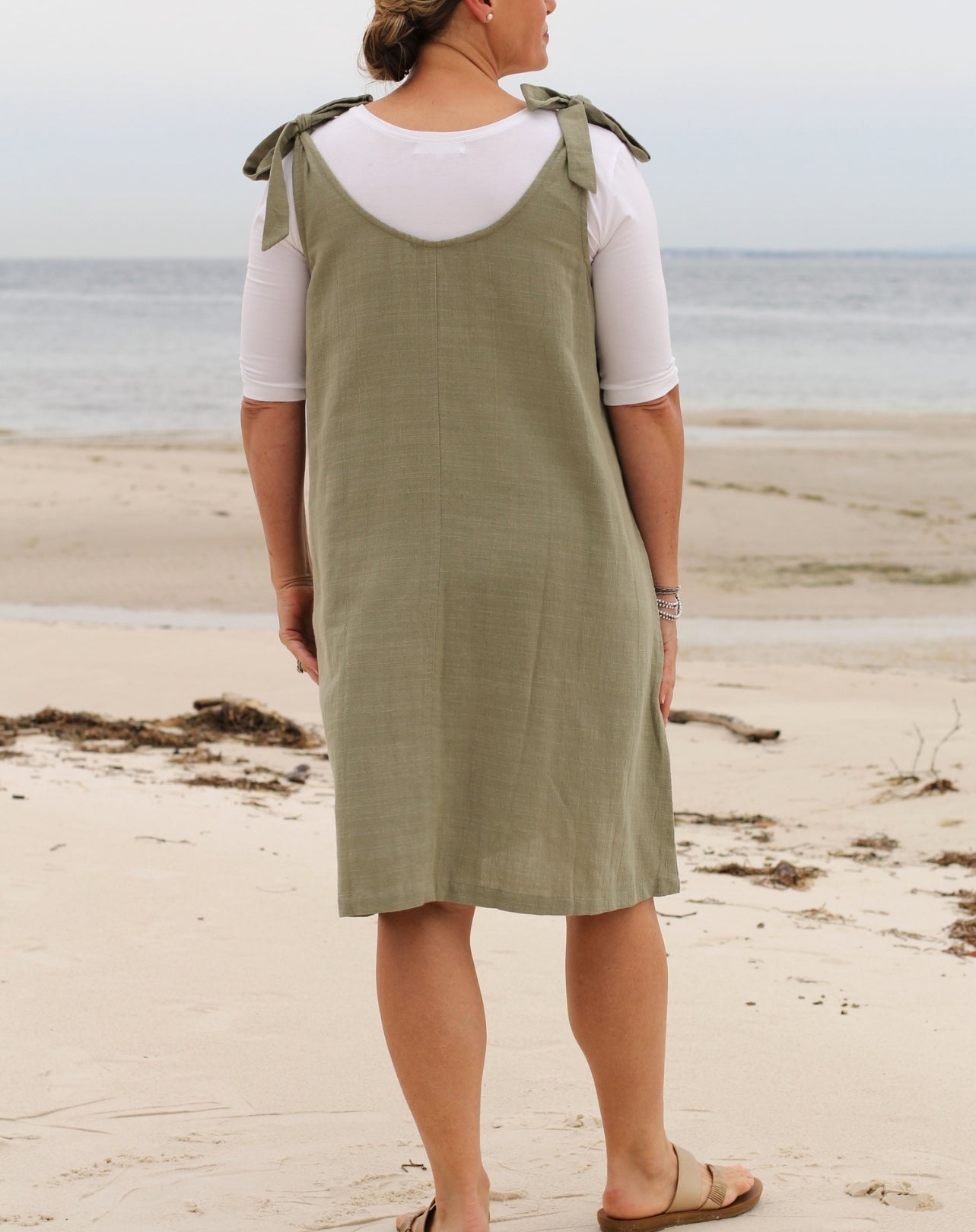 A woman standing on a sandy beach wearing a sleeveless green pinafore-style smock dress with ties over a white short-sleeve top, photographed from the back.