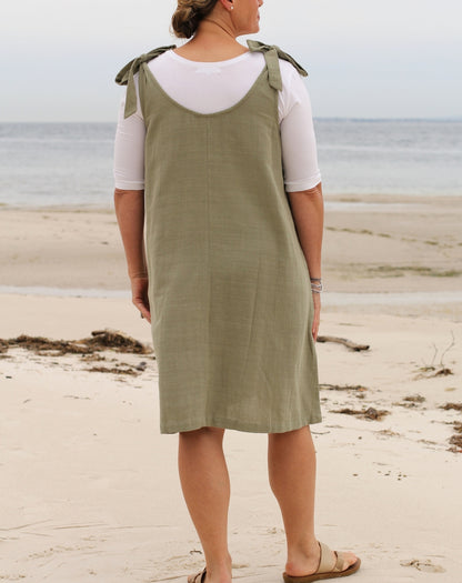A woman standing on a sandy beach wearing a sleeveless green pinafore-style smock dress with ties over a white short-sleeve top, photographed from the back.