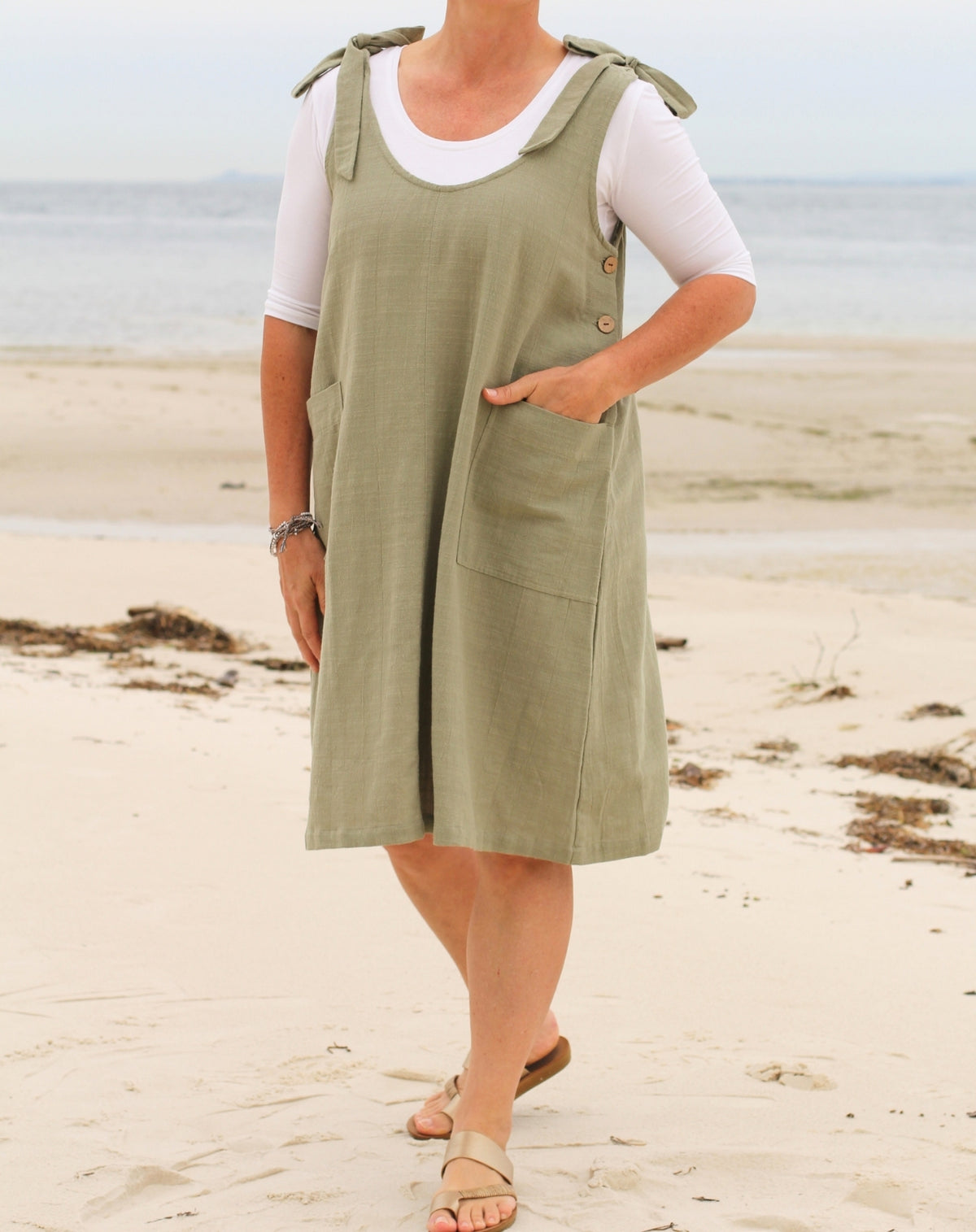 A woman wearing a sage green linen pinafore dress over a white t-shirt, standing on a sandy beach with the ocean in the background.