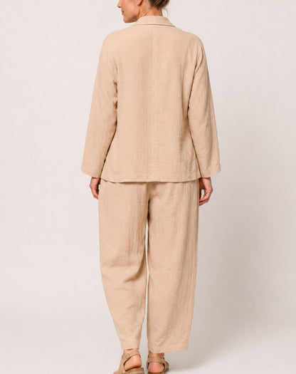A woman shown from the back wearing a light natural linen two-piece set consisting of a long-sleeve cotton blazer jacket and relaxed-leg trousers, paired with beige sandals against a plain light grey studio background.