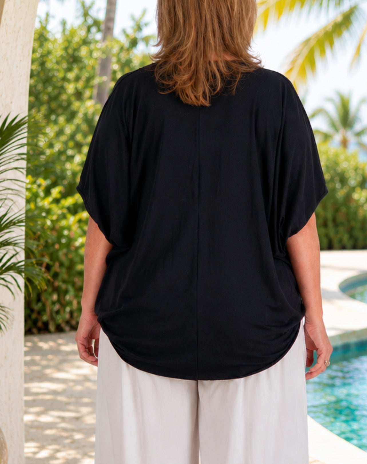A woman viewed from behind wearing a loose black short-sleeve top and white pants, standing outdoors near a pool with greenery in the background.