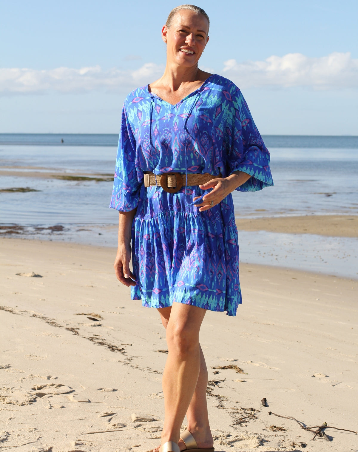 A midsized woman standing on a sandy beach wearing a blue and purple patterned boho-style dress with a brown belt, with the ocean and sky in the background.