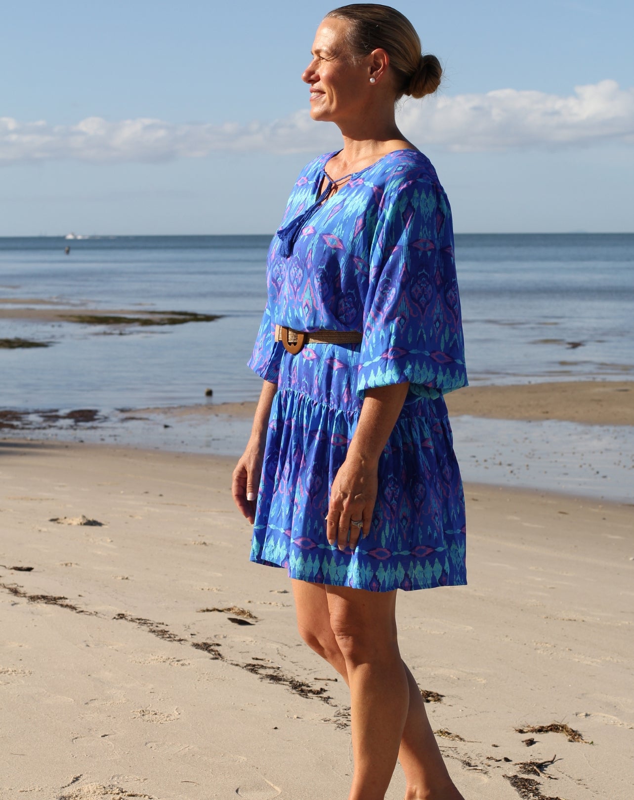 A woman wearing a blue and purple ikat patterned short boho dress with a brown belt walks along a sandy beach with the ocean in the background.