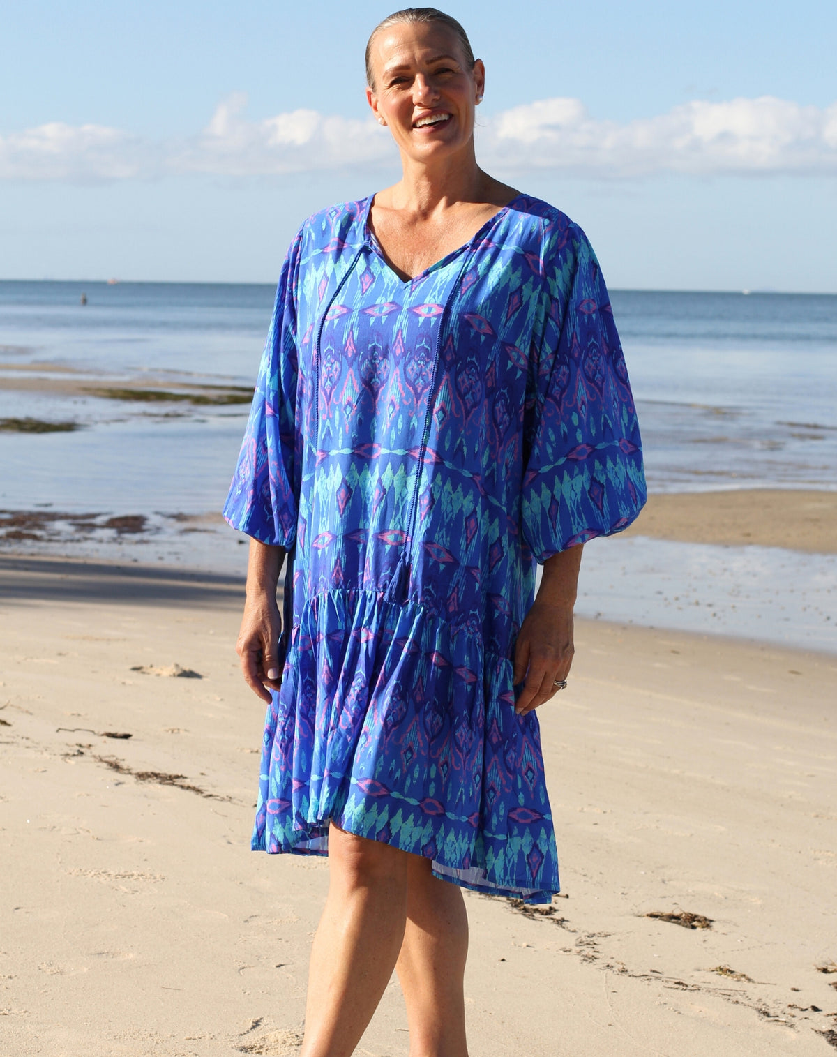A mature woman wearing a blue and purple ikat patterned boho shift dress stands on a sandy beach with the ocean and sky in the background.